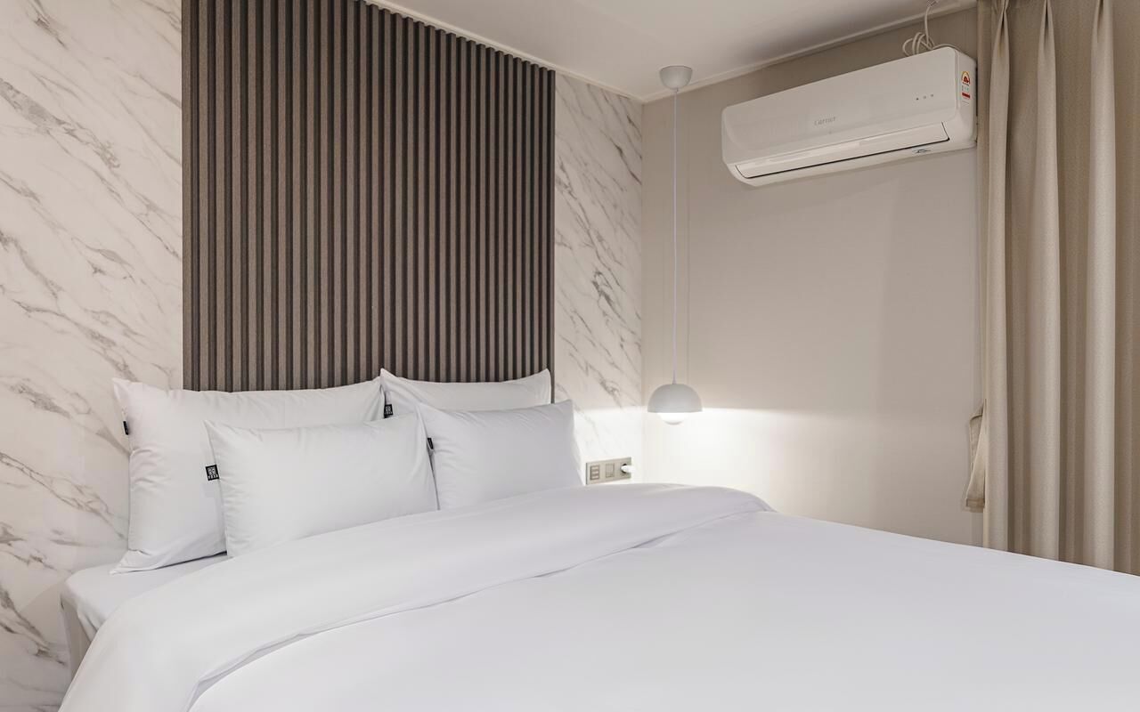 Una habitación de hotel con cama, ropa de cama blanca, aire acondicionado de pared y una lámpara de noche.