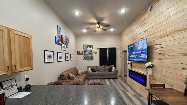 A modern kitchen with granite countertops, wooden cabinets, a dining area with a table and chairs, a TV mounted on the wall, and a ceiling fan.