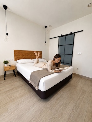 A modern bedroom with a wooden headboard, a bedside table with a plant, and a large window with a view. - Apartamento cómodo cerca del Puerto  (València)