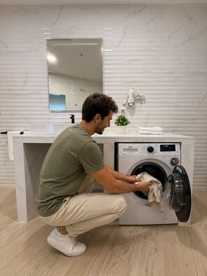 A person kneeling in front of a washing machine in a modern bathroom with a marble wall and a sink. - Apartamento cómodo cerca del Puerto  (València)