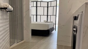 A modern hotel room with a glass shower enclosure, a bed with a black headboard, and a built-in washing machine.