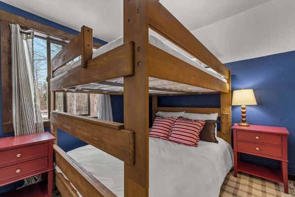 A bunk bed with a wooden frame, a red nightstand with drawers, a bedside lamp, and a window with curtains.