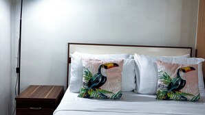 A neatly made bed with white bedding and two decorative pillows featuring a toucan design, a bedside table with a lamp, and a wooden nightstand.