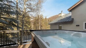 Ett bubbelbad på ett trädäck med utsikt över träd och ett hus i bakgrunden.