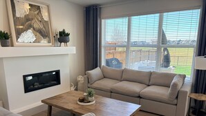 A living room with a sofa, a coffee table, a fireplace, and a large window with a view of a residential area. - Spacious 5BR Retreat w/ Hot Tub, Fire Pit, Gym & Movie Room (Calgary)