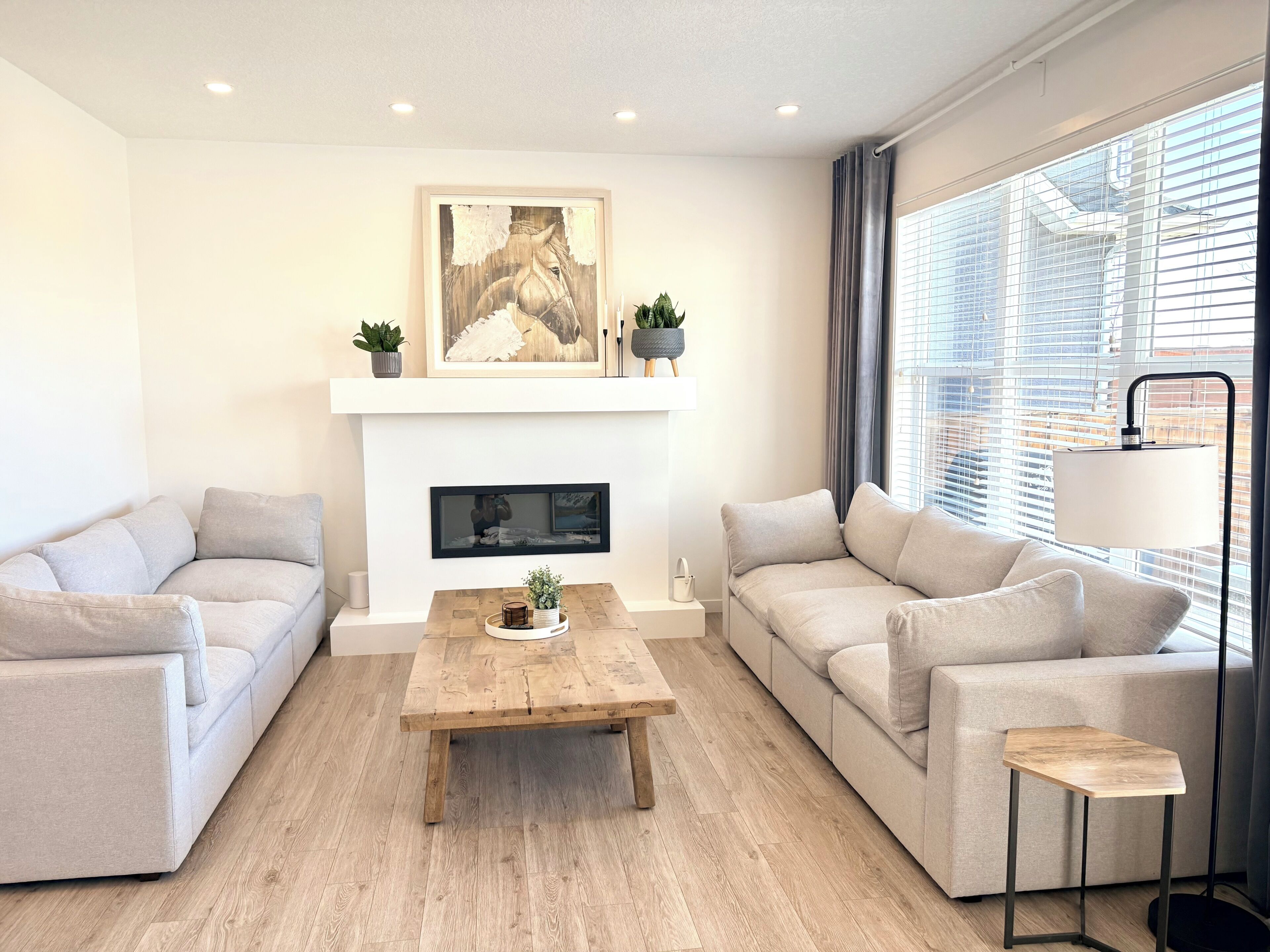 Ein modernes Wohnzimmer mit Kamin, zwei Sofas, einem Couchtisch und einem großen Fenster mit Jalousien.