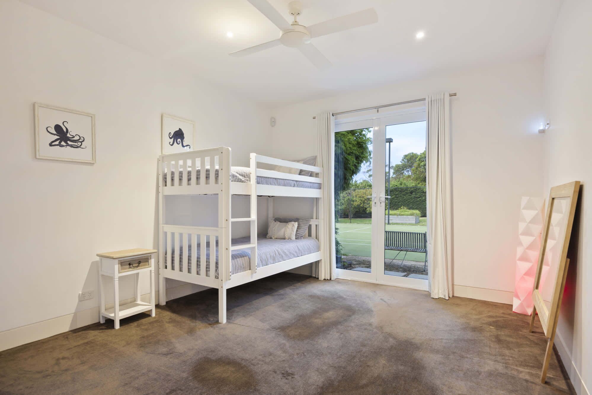 A room with a bunk bed, a small bedside table, a ceiling fan, and a view of a tennis court through a glass door.