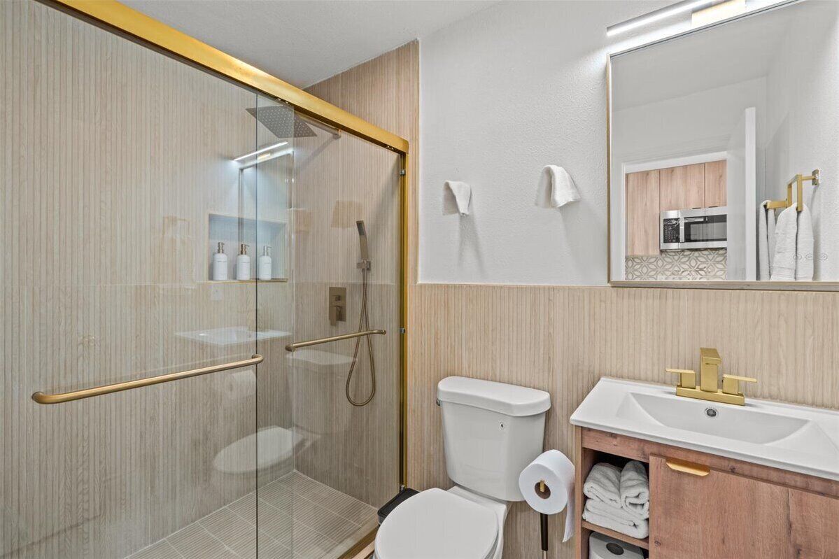Un baño moderno con cabina de ducha de vidrio, inodoro, lavamanos con grifo dorado y espejo sobre el lavamanos.