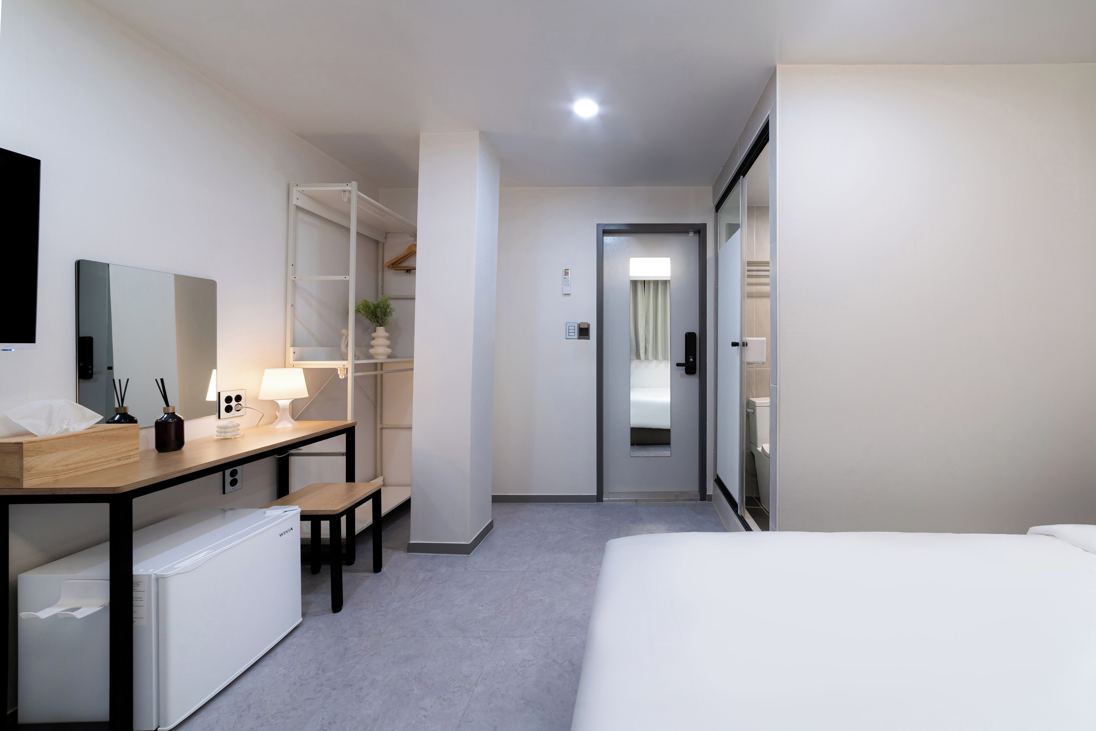 Een hotelkamer met een bed, een bureau met lamp, een kleine koelkast, een spiegel en een garderobe.