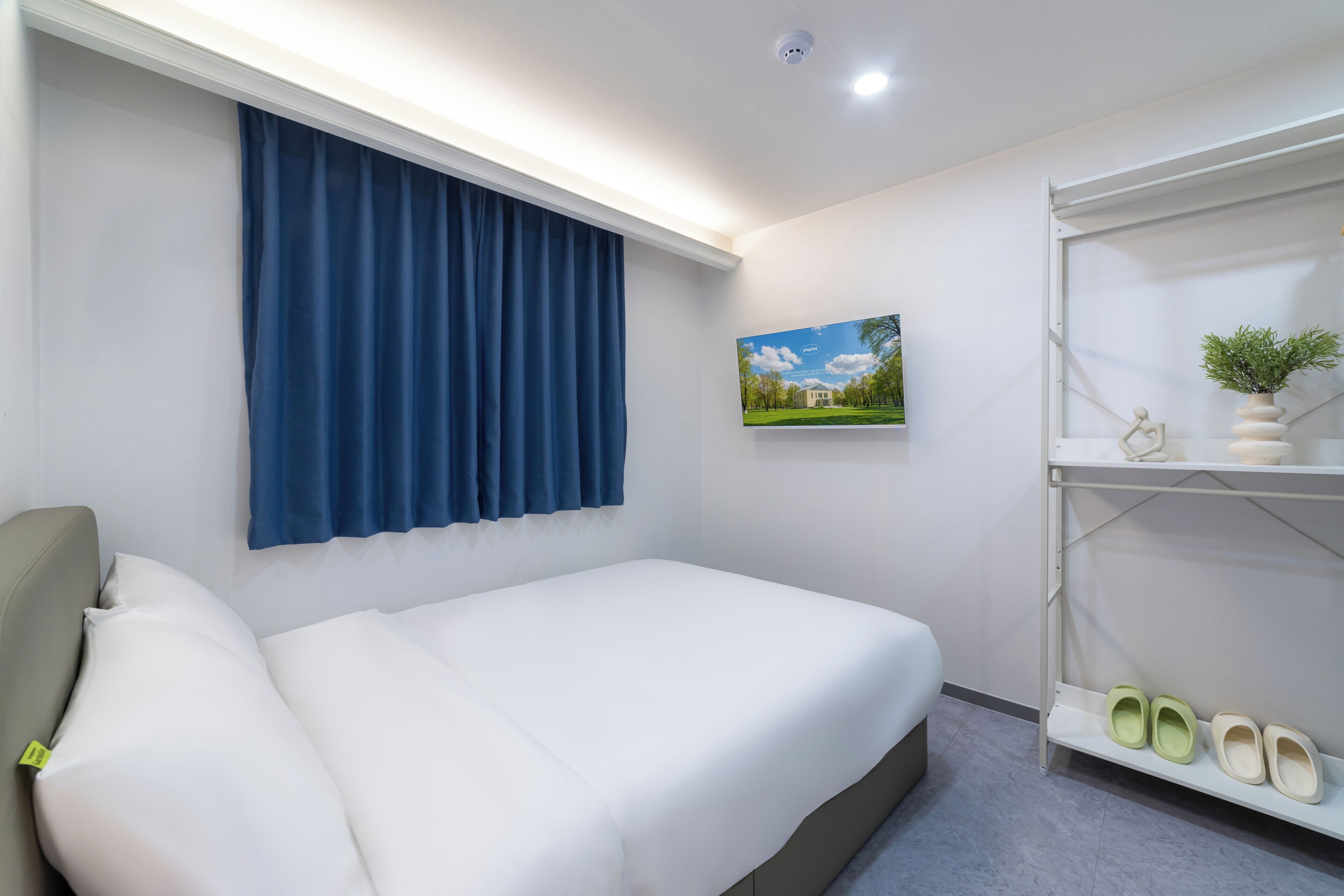 A hotel room with a bed, a blue curtain, a picture on the wall, and a shelf with shoes.