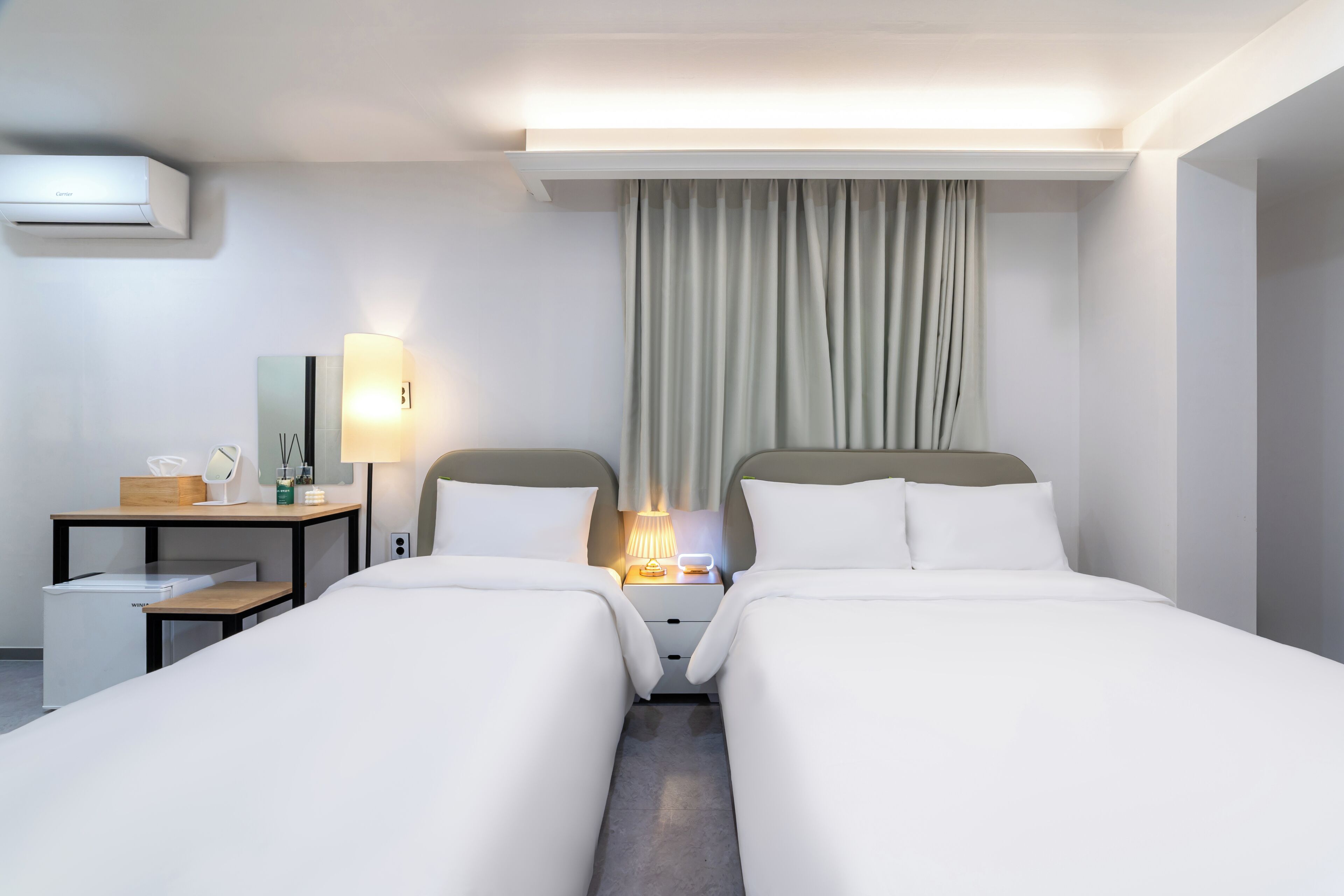 Quarto de hotel com duas camas, uma escrivaninha, uma luminária e um ar condicionado.