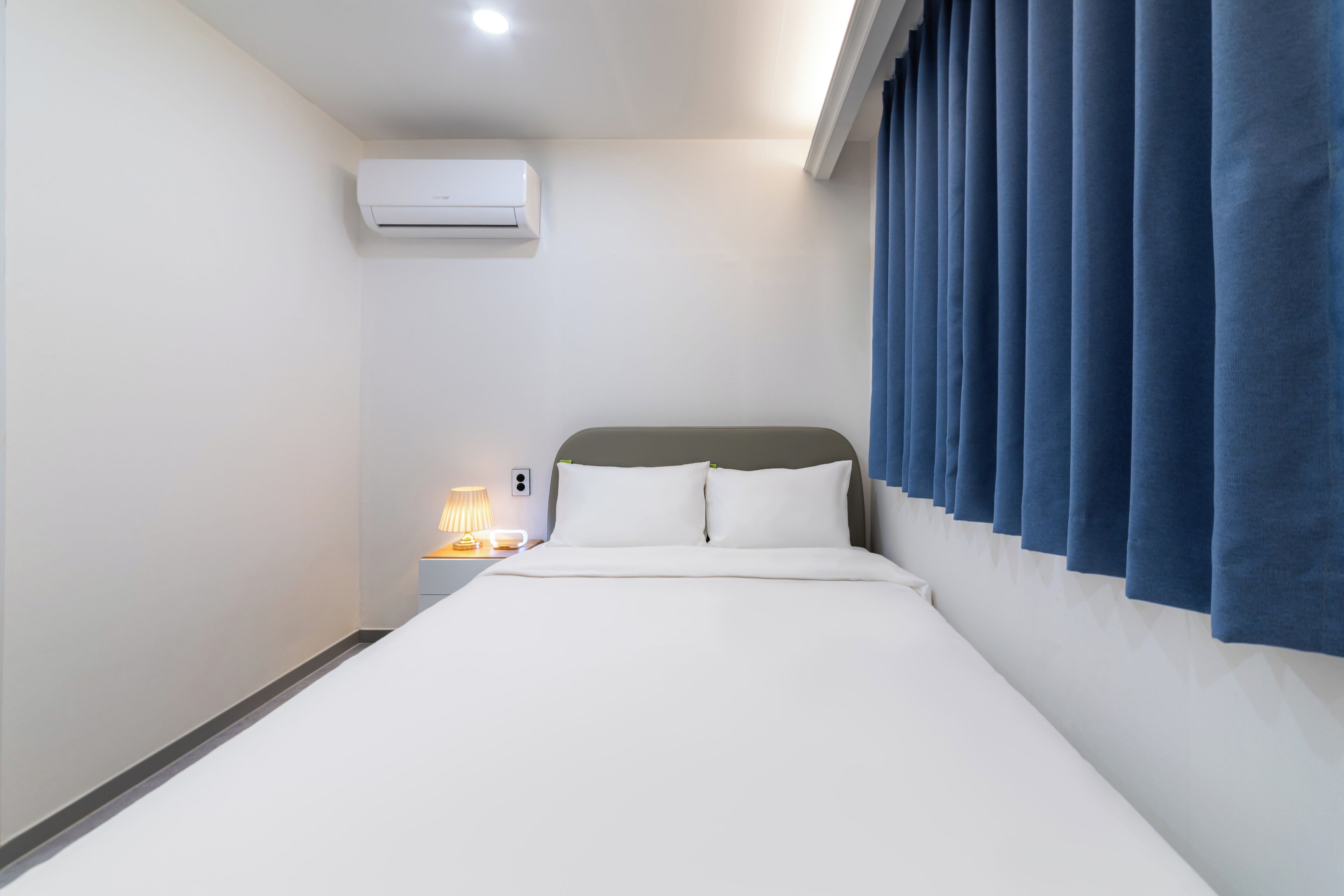 A hotel room with a bed, an air conditioner, a nightstand with a lamp, and blue curtains.