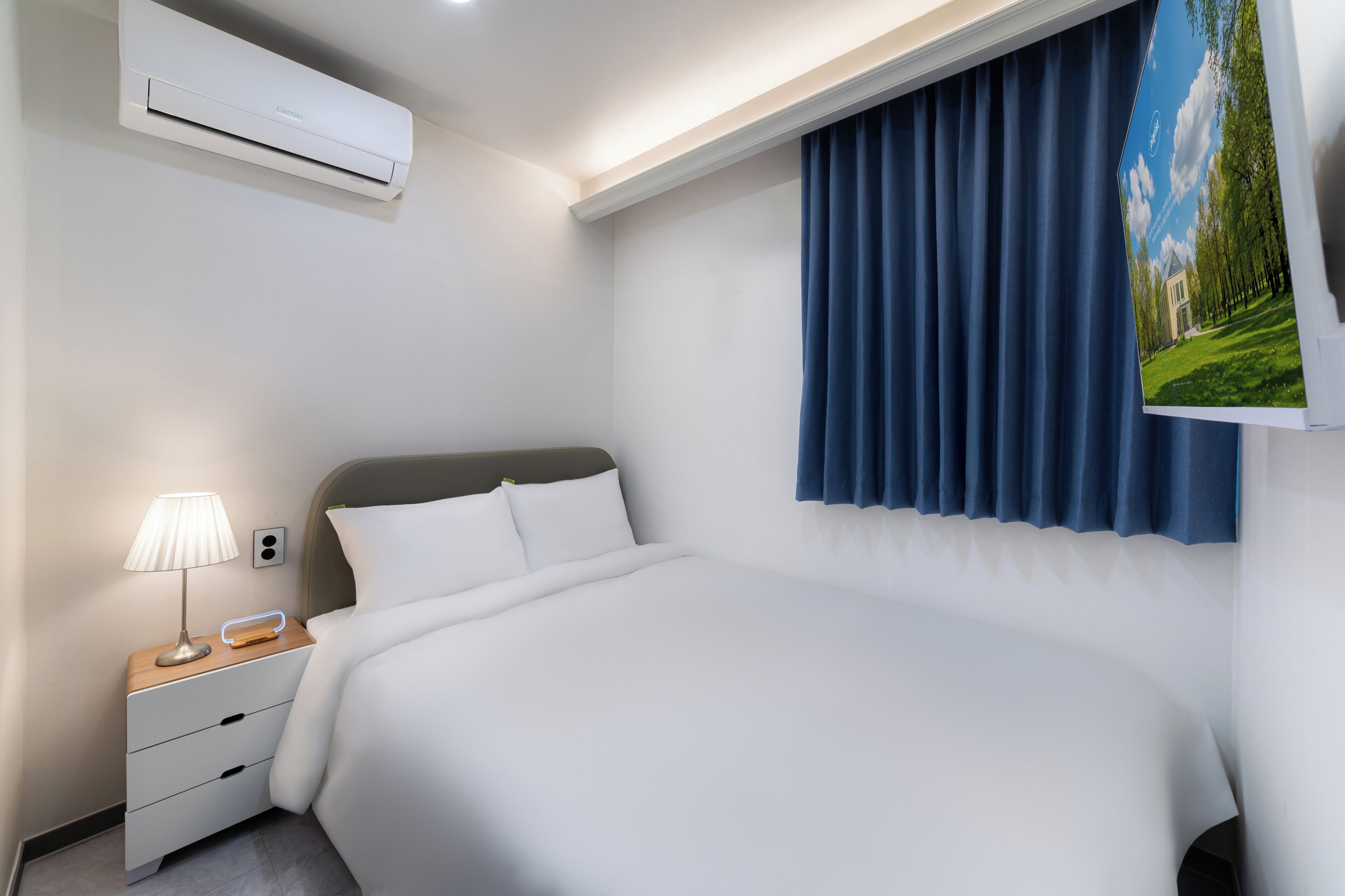 A hotel room with a bed, a nightstand with a lamp, an air conditioner, and a window with curtains.