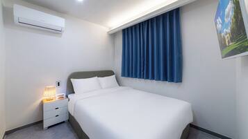 Una camera d'albergo con un letto, un comodino con una lampada, un condizionatore d'aria e una finestra con tende.