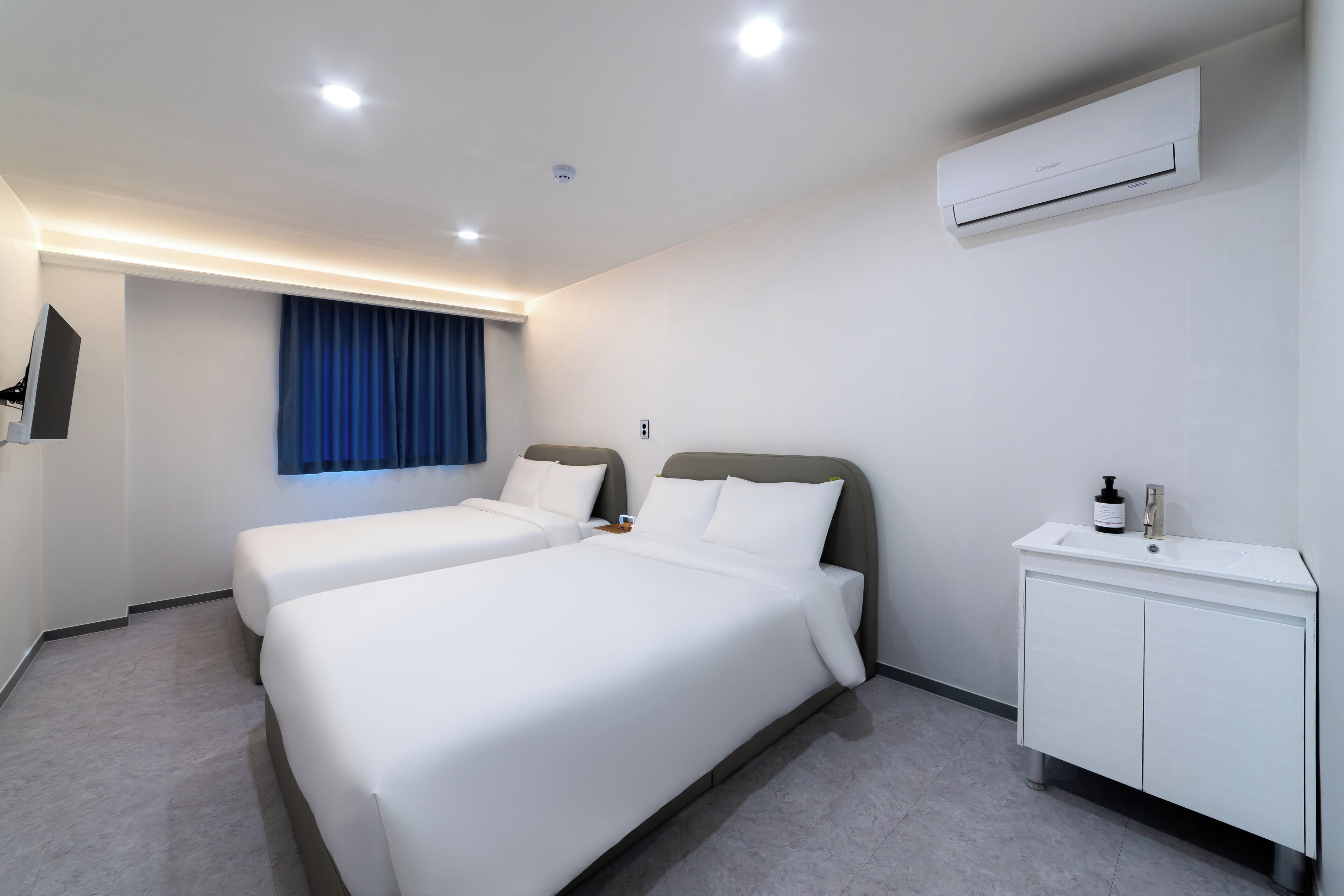 A hotel room with two beds, a white vanity with a sink, and an air conditioning unit on the wall.
