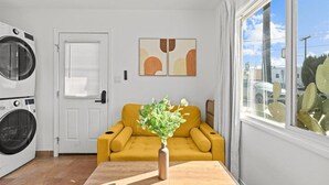 Ein gemütlicher Wohnraum mit Waschmaschine, einem gelben Sofa, einer Vase mit Grünpflanzen und abstrakter Wandkunst.