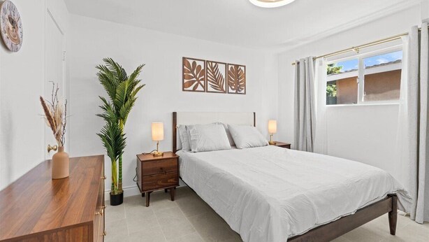 Ein Schlafzimmer mit Bett, Nachttischen, einer Zimmerpflanze, Wandbildern und einem Fenster mit Vorhängen.