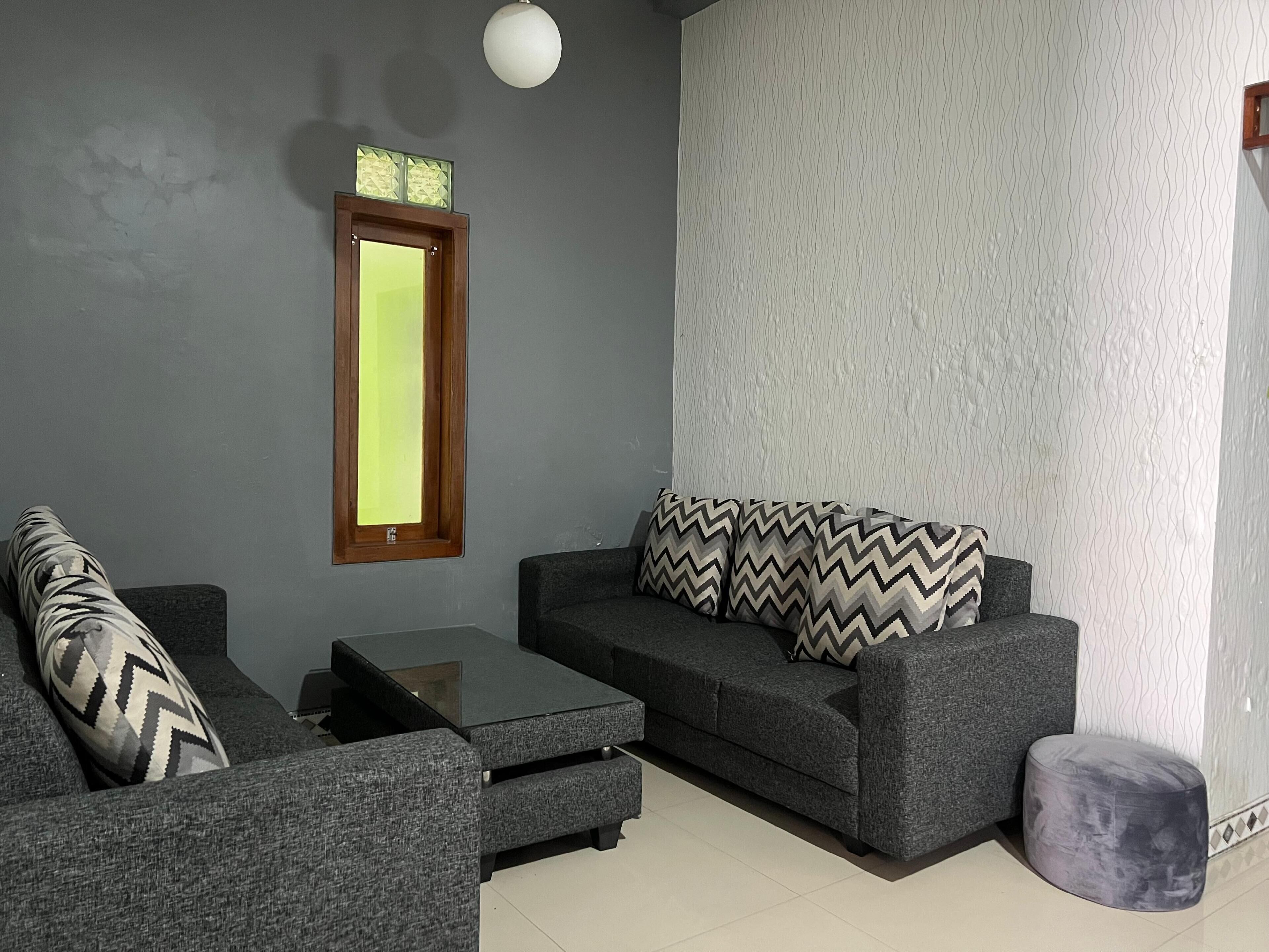 Ein modernes Wohnzimmer mit einem grauen Sofa, einem passenden Hocker und einem Couchtisch. Über dem Sofa hängt ein Spiegel und die Wand ist mit einem Muster versehen.