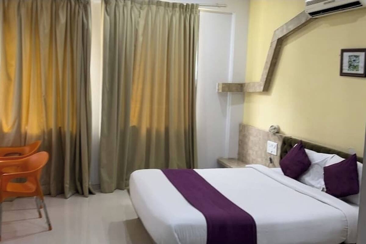 A hotel room with a bed, curtains, an air conditioning unit, a chair, and a small table.