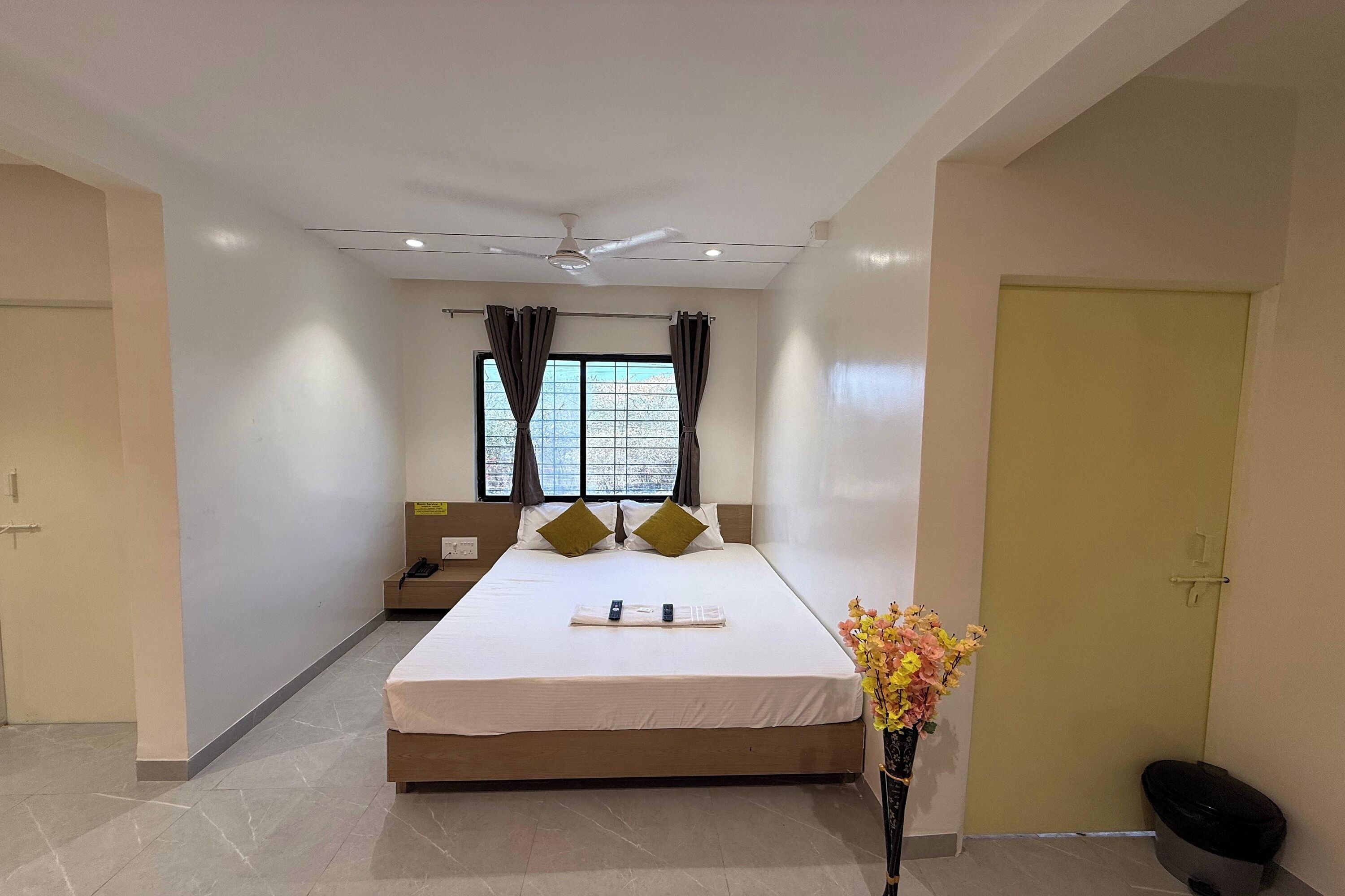 A hotel room with a bed, a bedside table, a window with curtains, a ceiling fan, and a door.