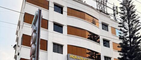 Un edificio de varios pisos, un letrero que dice 'INDRAPRASTHA LODGING' y un letrero que anuncia 'HABITACIONES PREMIUM a partir de 499 Rs.'.