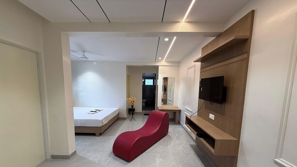 Una habitación de hotel moderna con una butaca roja, una cama, un televisor montado en un estante de madera y un ventilador de techo.