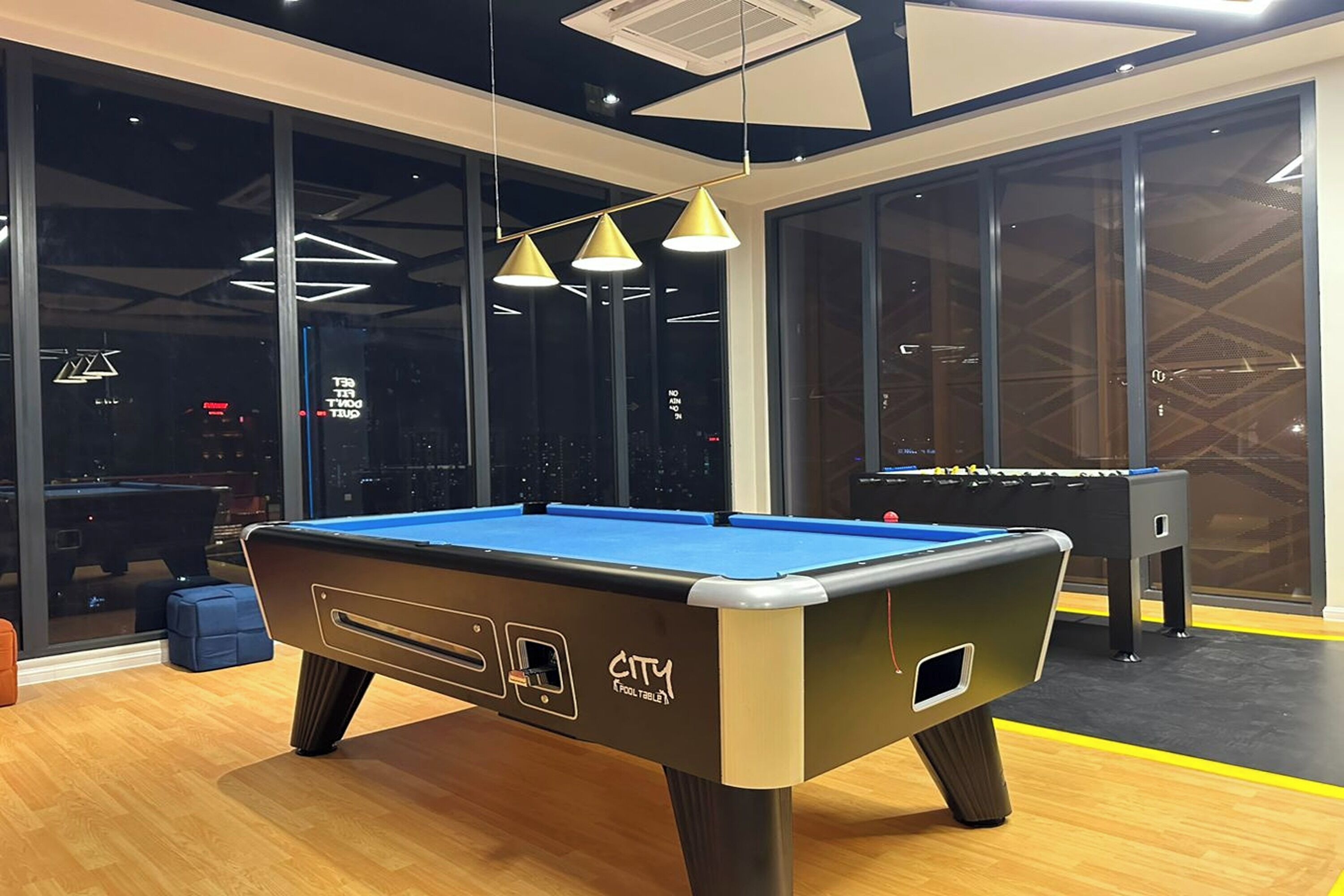 A pool table with a CITY logo, a foosball table, and a window with a city view.