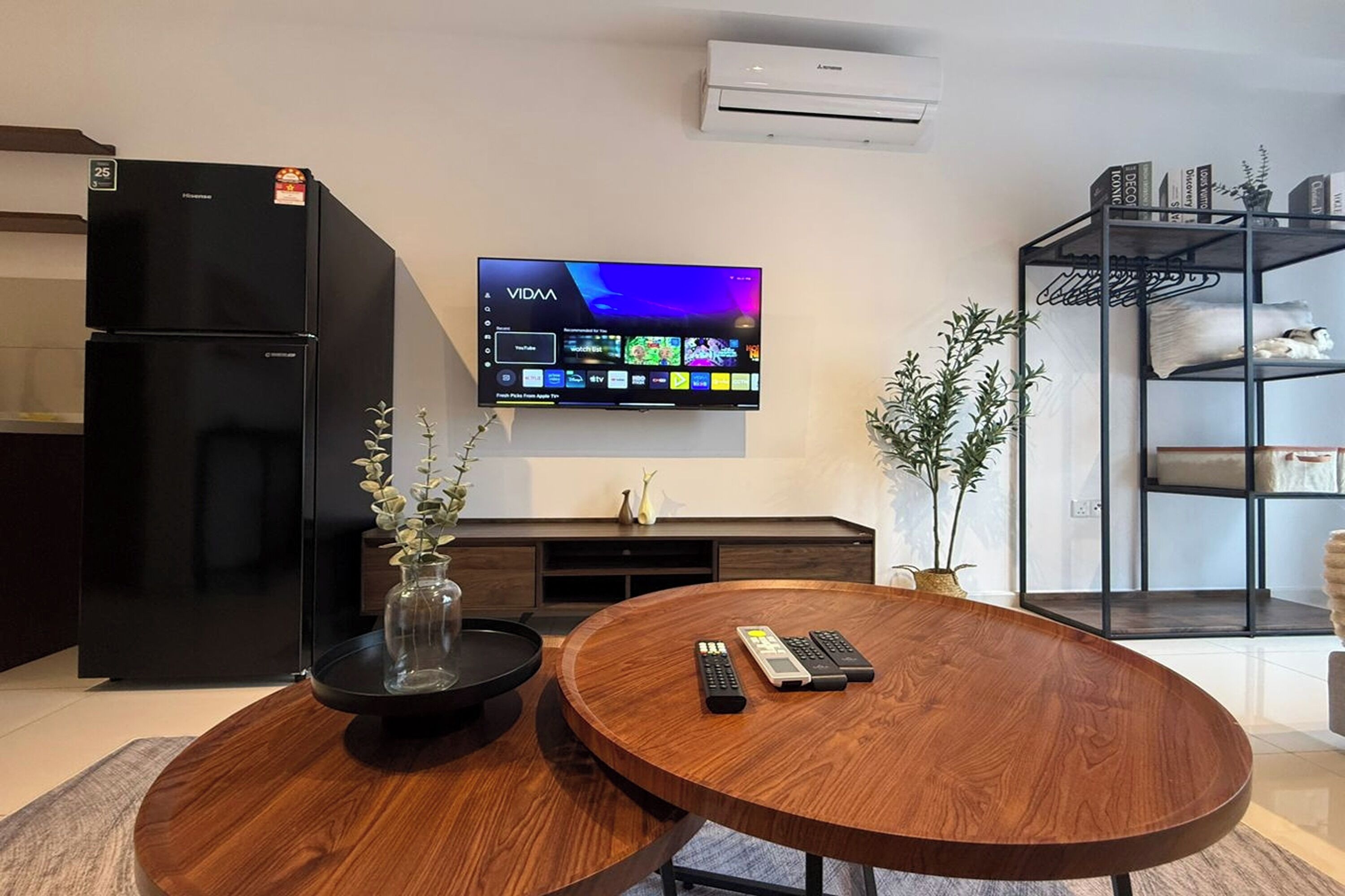 A modern living room with a flat-screen TV mounted on the wall, a black refrigerator, a wooden coffee table, and a bookshelf with decorative items.