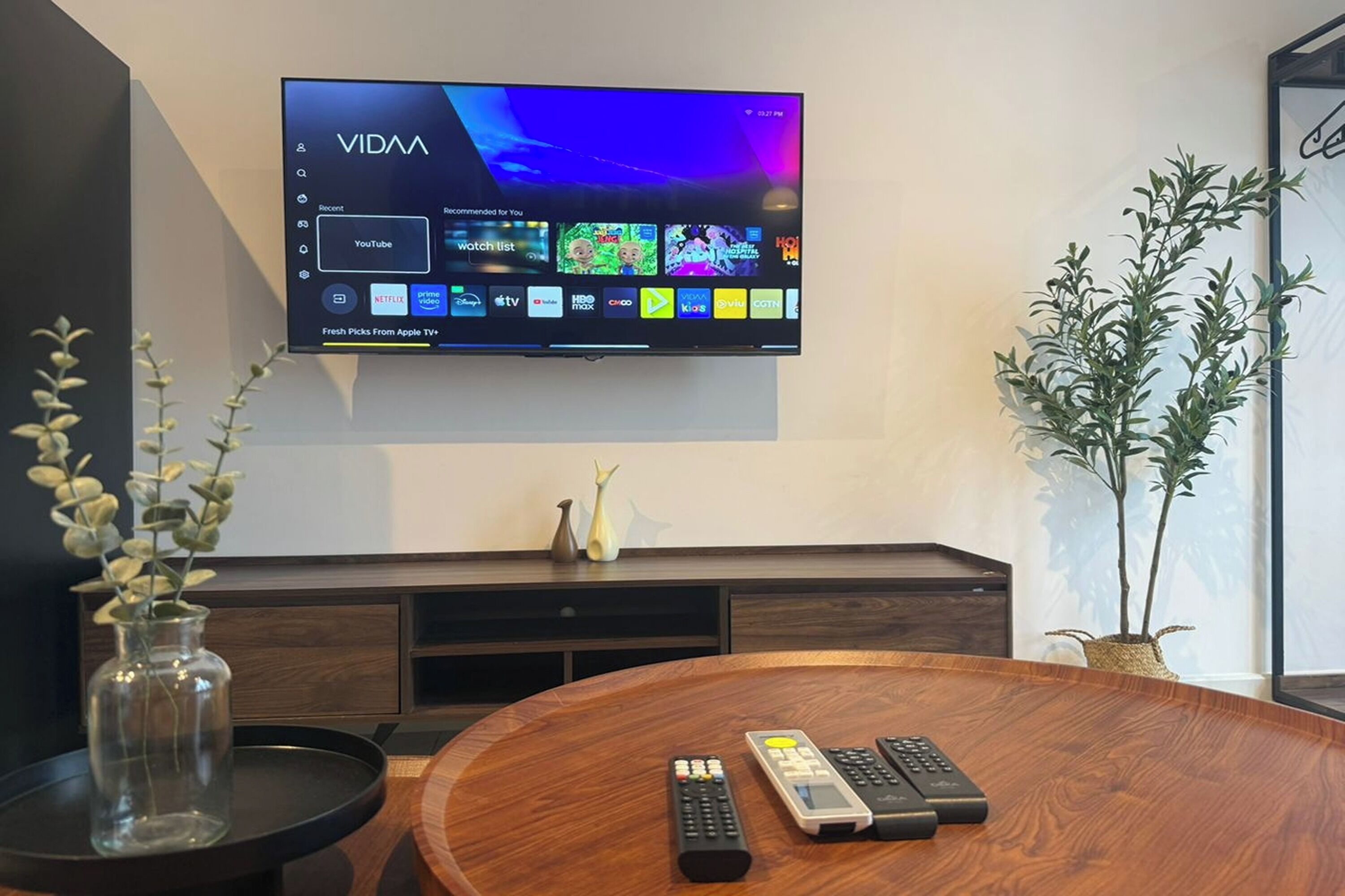 A modern living room with a mounted TV displaying a streaming service interface, a wooden coffee table with remote controls, a vase with greenery, and a potted plant.