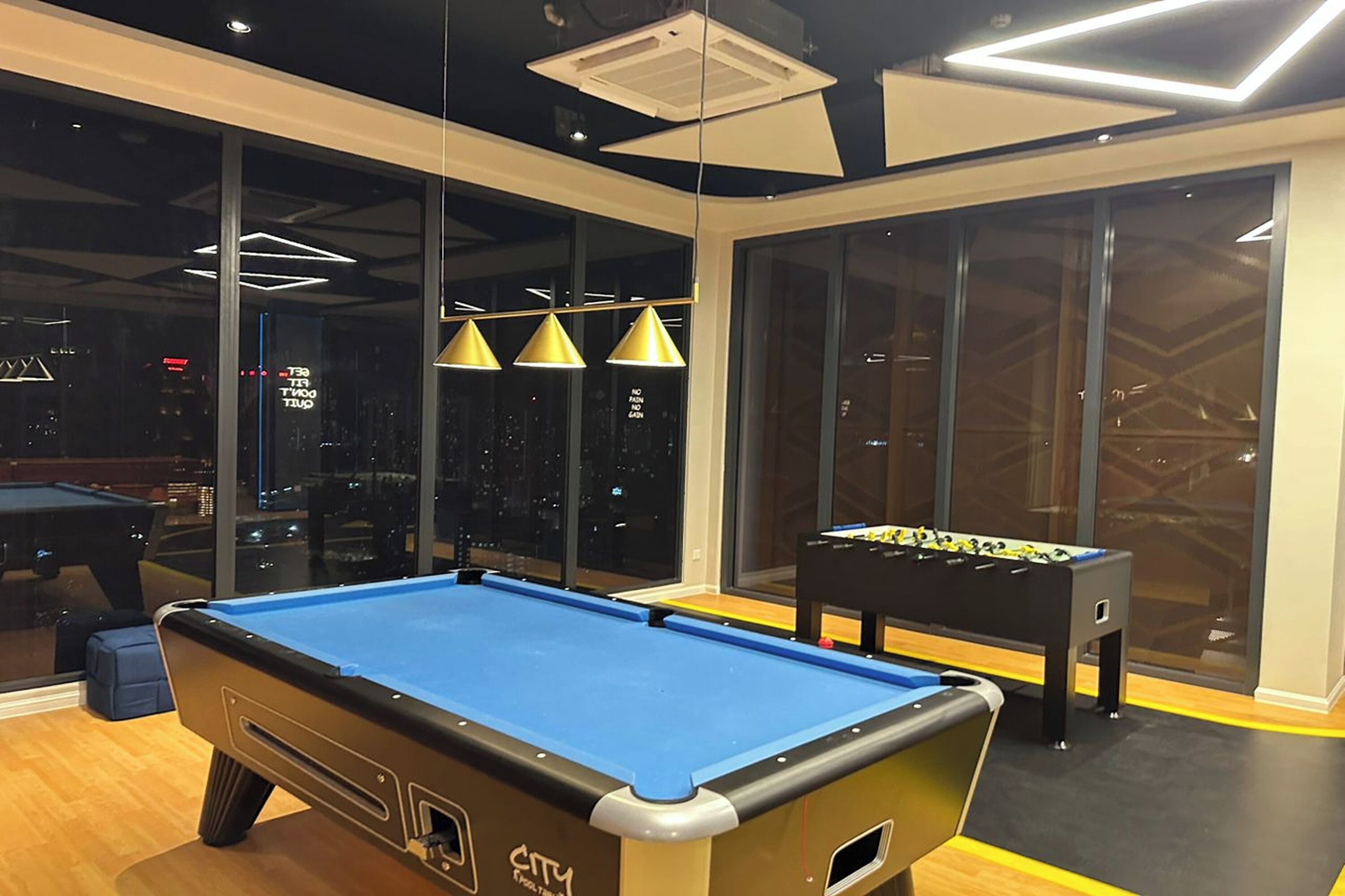 A pool table with a blue felt surface, a foosball table, and pendant lighting in a room with large windows.