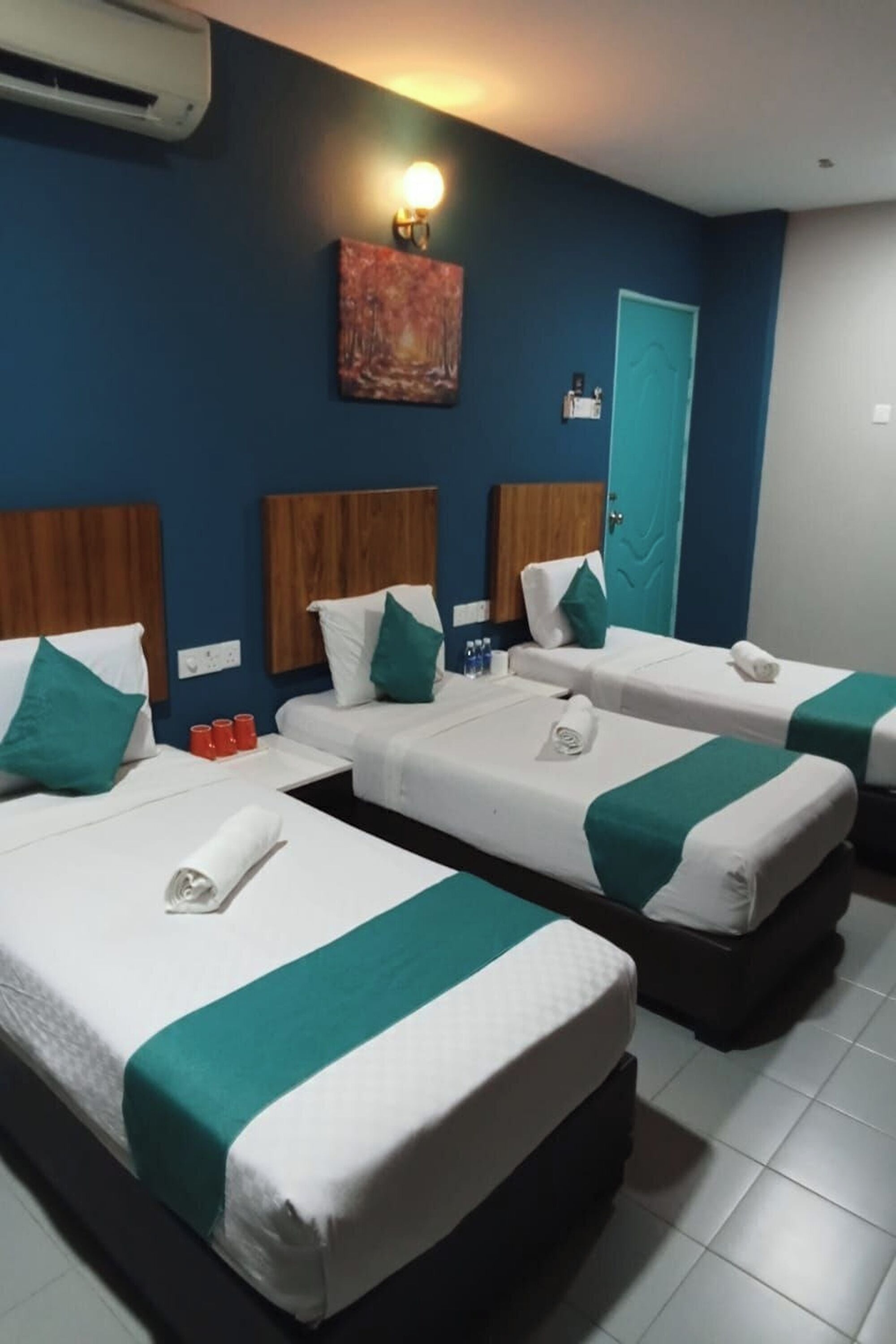Habitación de hotel con tres camas individuales, cada una con cubrecama color turquesa y ropa de cama blanca, cabecero de madera y aire acondicionado instalado en la pared.