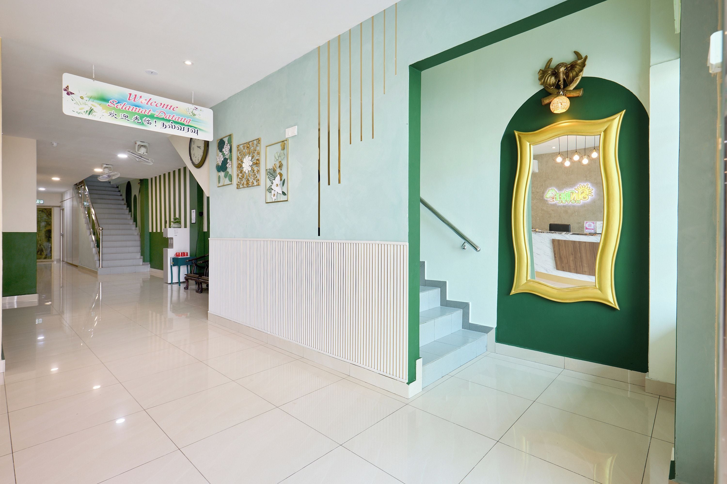 A hotel reception area with a green wall, a reception desk, a staircase, and a sign welcoming guests.