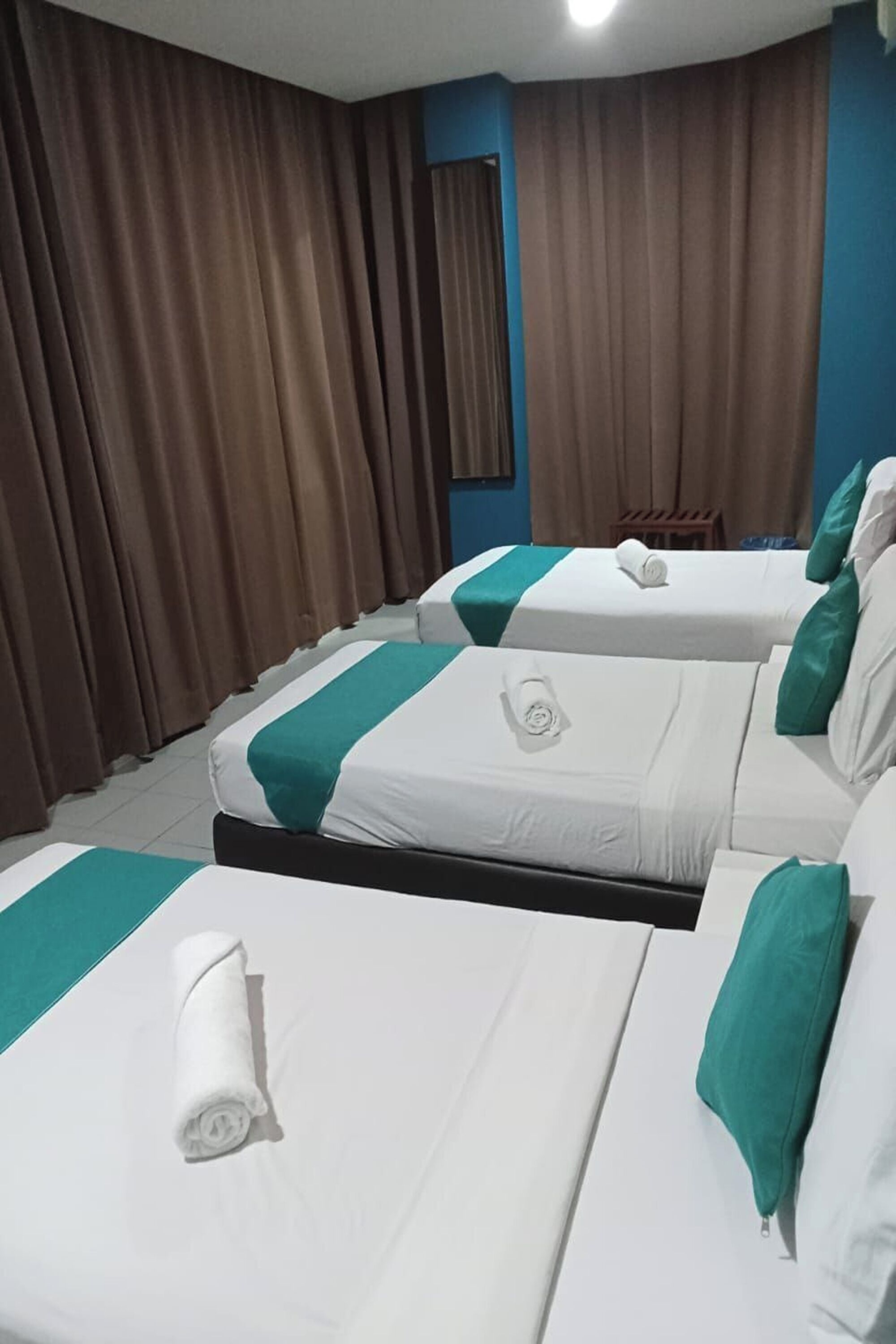 A hotel room with three beds, each with white bedding and teal accents, brown curtains, and a small wooden stool.