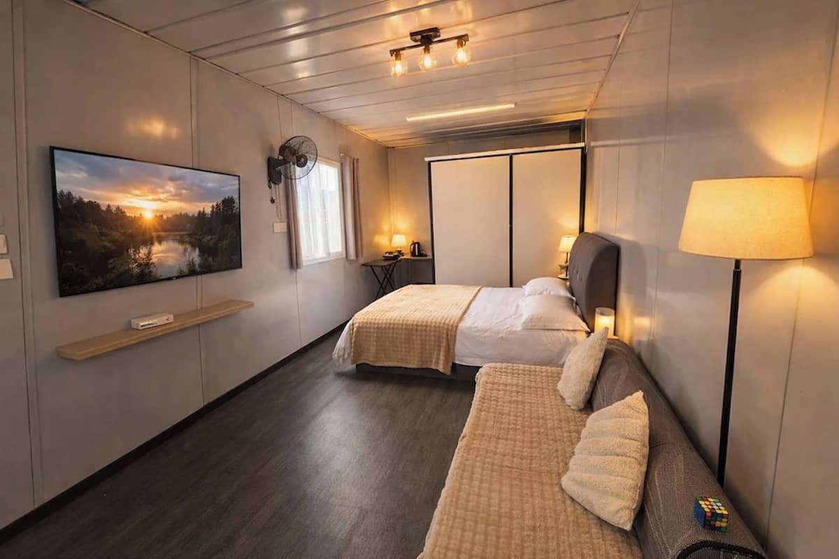 A hotel room with two beds, a TV, a fan, a lamp, and a view of a sunset.