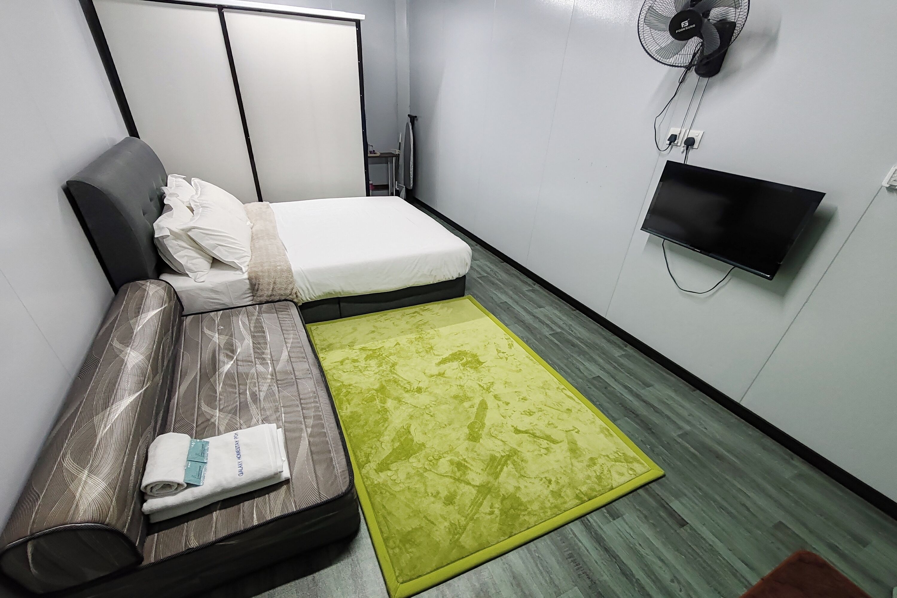 Ett litet hotellrum med en säng, en soffa, en tv, en fläkt och en grön matta.