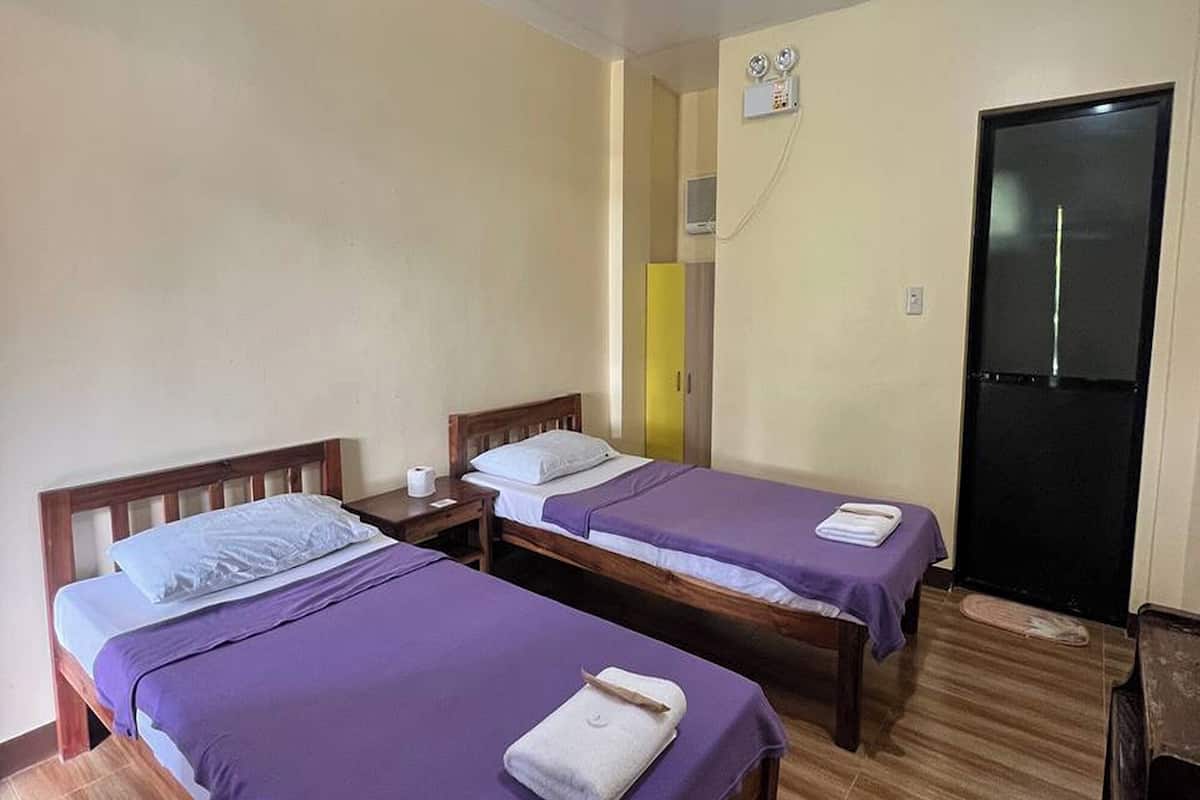 Egy szobában két egyágyas ágy található, melyeknek fa keretük és lila ágyneműjük van. A szobában fa padló és fekete ajtó is van.