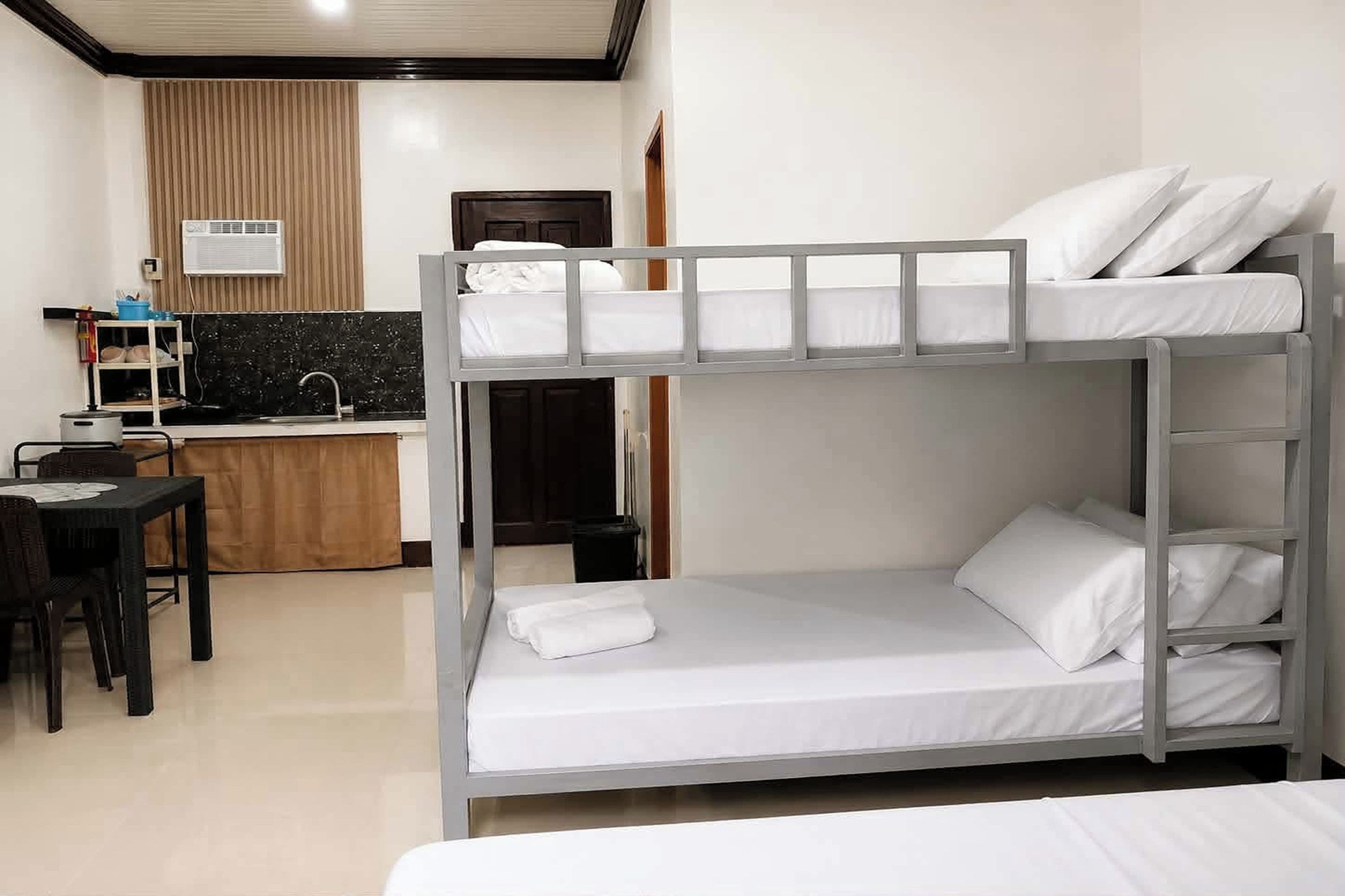 A bunk bed with white bedding, a small desk with a chair, and a kitchenette with a sink and stove.
