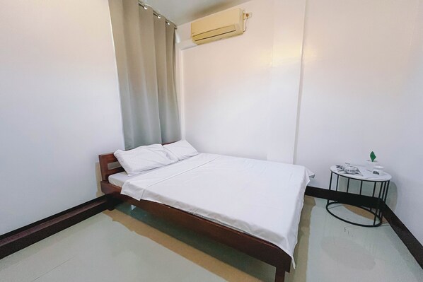 Un lit simple avec un cadre en bois, un matelas blanc et un oreiller blanc. Il y a une petite table ronde avec un plateau blanc et un pied noir, ainsi qu’un appareil de climatisation fixé au mur.
