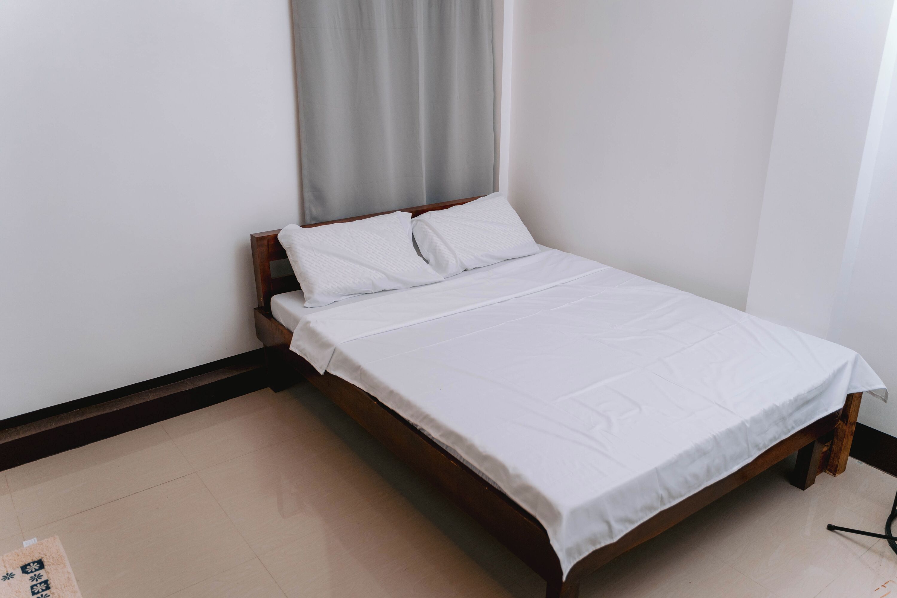 A single bed with a wooden frame and white bedding in a room with plain walls and a tiled floor.