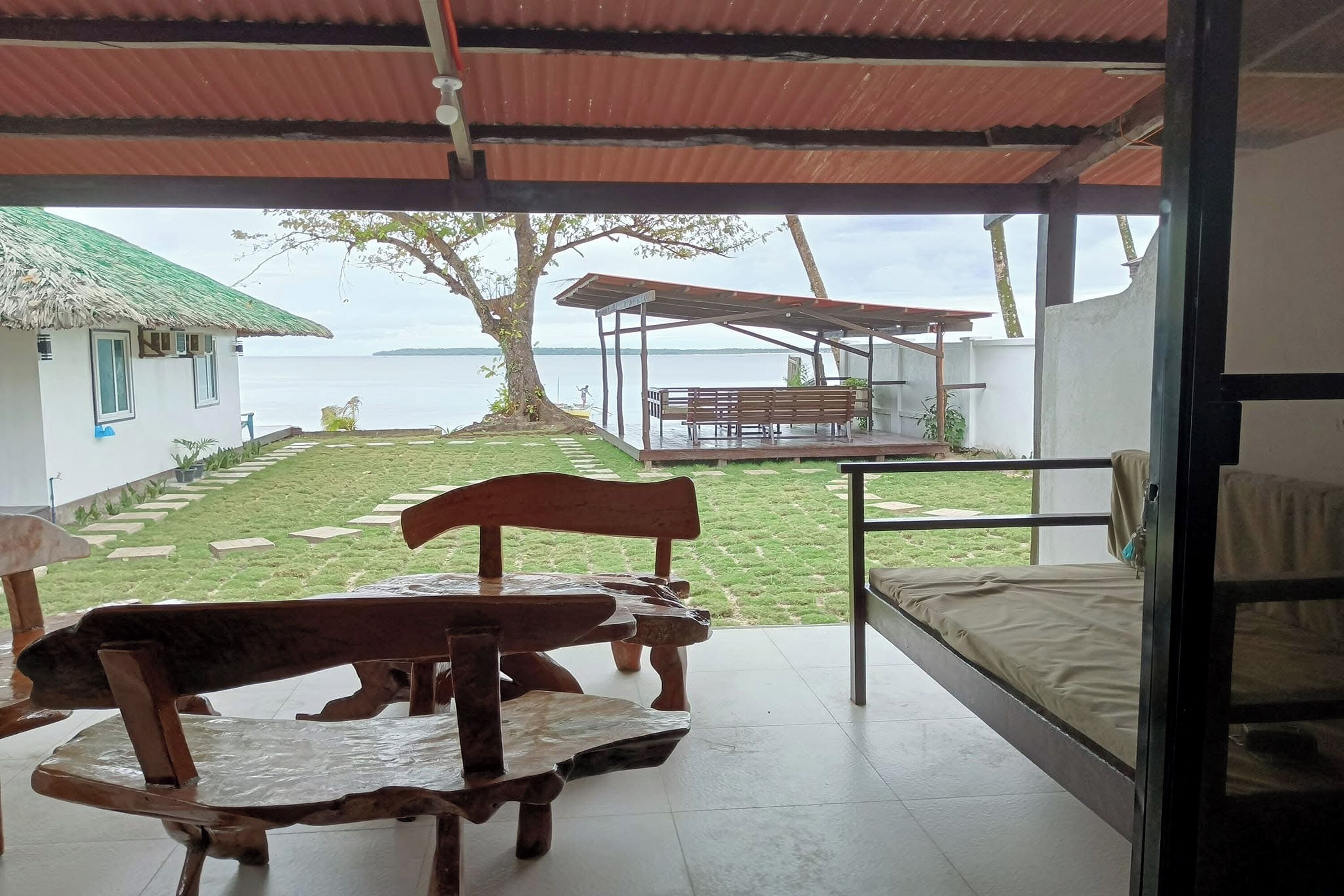 A covered outdoor seating area with a table and benches, a bed, and a view of a body of water and a grassy area with a tree.