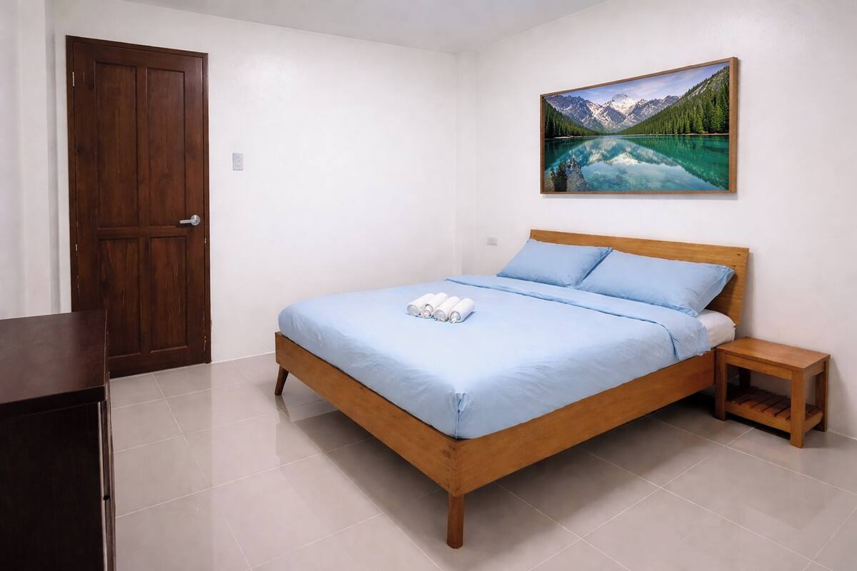 A bedroom with a wooden bed, a small bedside table, a door, and a painting of a mountain landscape.