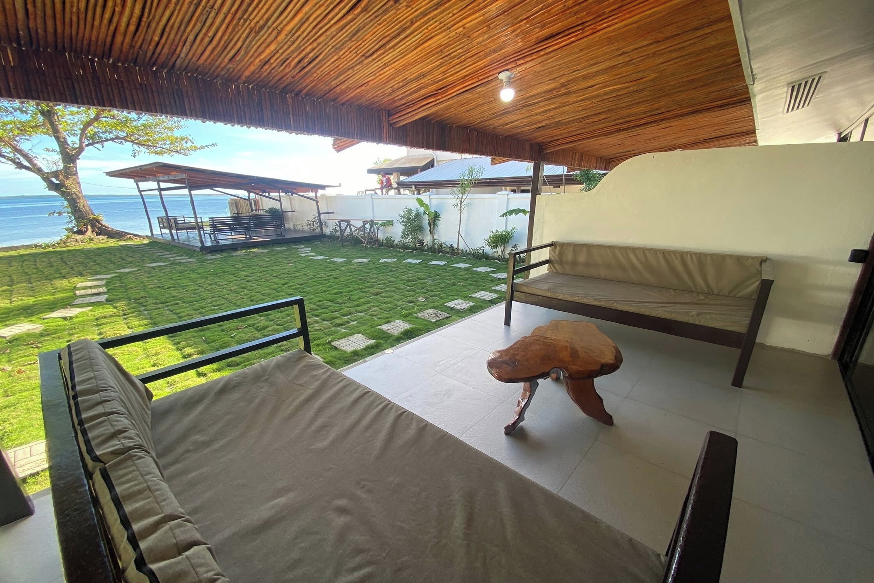 A covered outdoor seating area with a view of the sea, a grassy lawn, and a tiled patio.