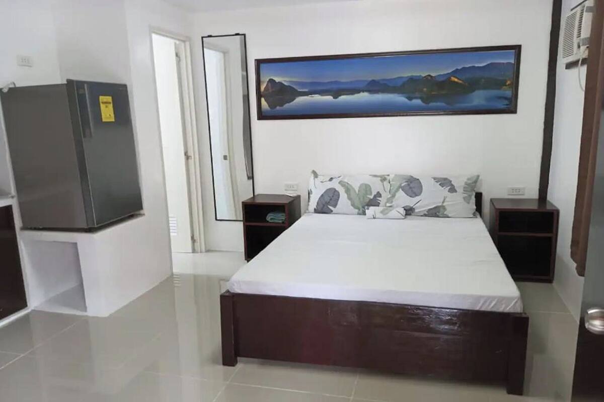 A bedroom with a bed, a nightstand, a bedside table, a refrigerator, and a door.