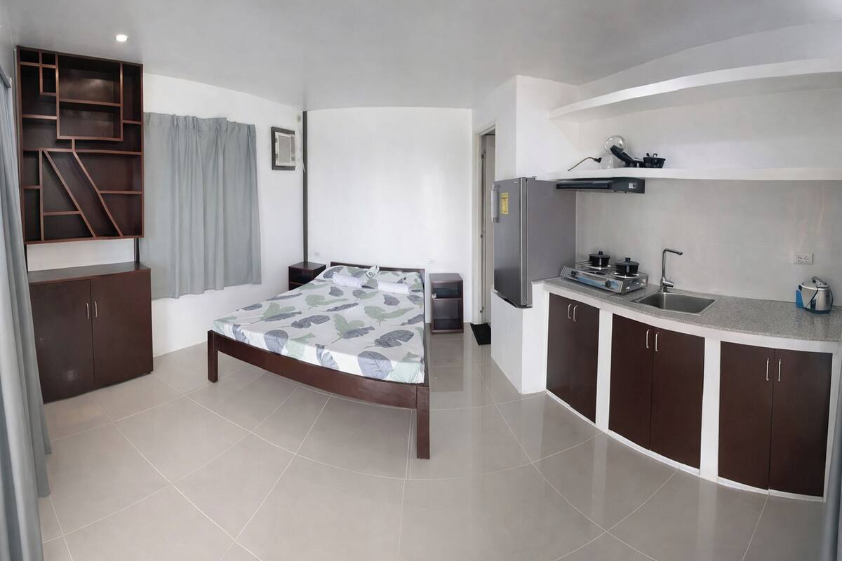 A bedroom with a bed, a built-in wardrobe, a kitchenette with a refrigerator, and a window with curtains.