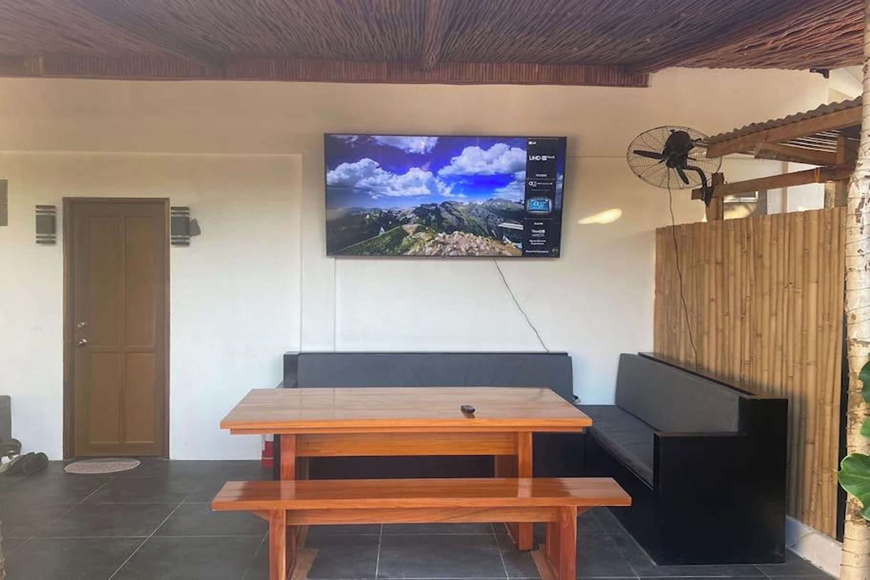 A room with a wooden table, a bench, a TV mounted on the wall, and a fan.