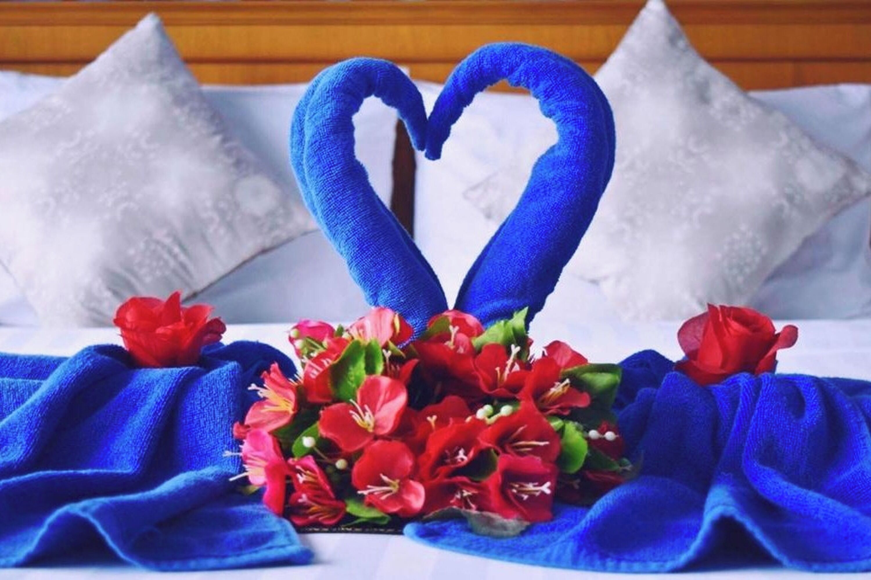 A bed with a blue blanket, a heart-shaped towel arrangement, and a bouquet of red flowers.