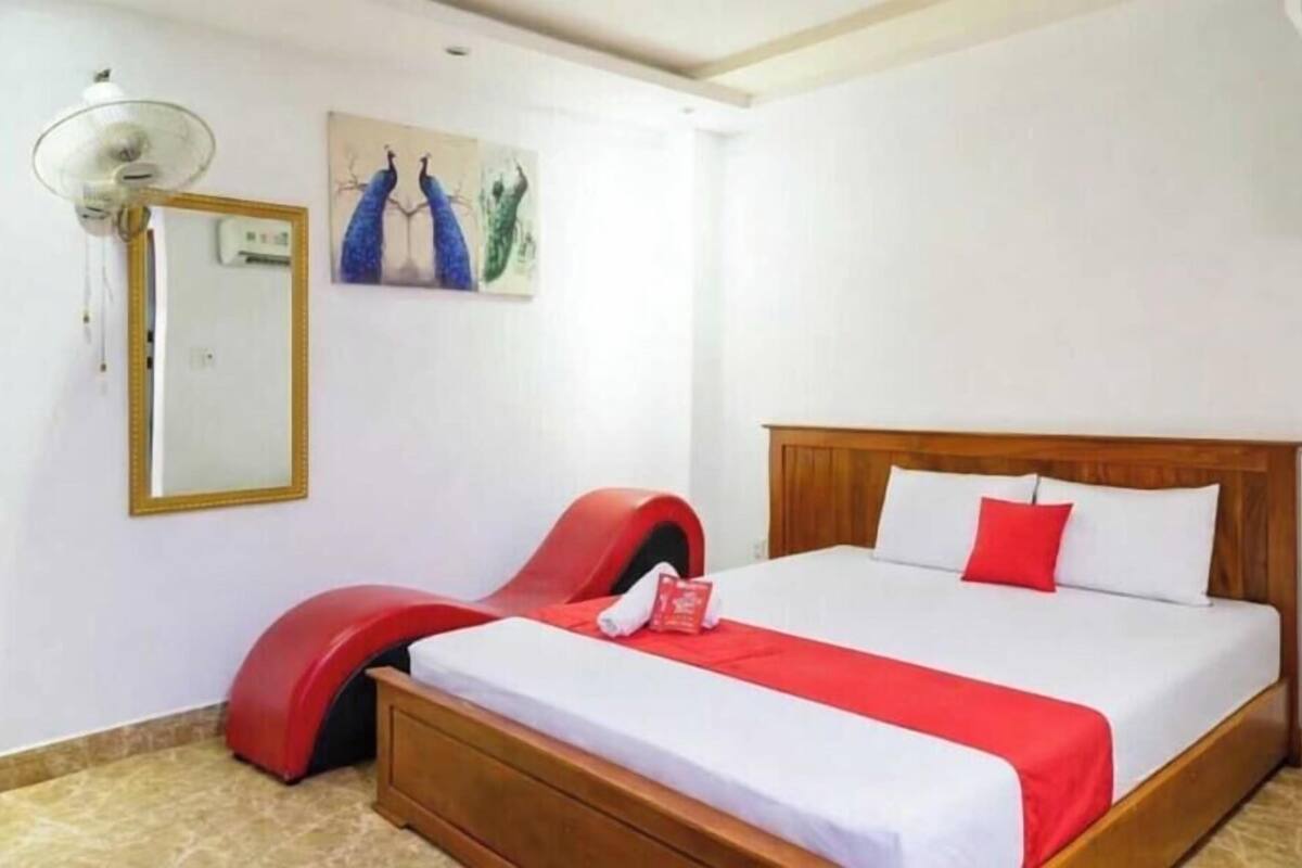 Une chambre d’hôtel avec un lit en bois, une chaise rouge, un ventilateur de plafond et un tableau accroché au mur.
