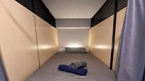 Une petite pièce étroite, avec un lit simple, un oreiller et une serviette enroulée.
