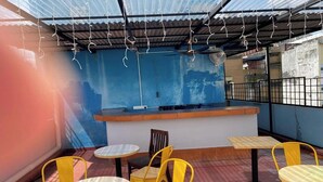 Un espace extérieur aménagé avec des chaises jaunes et des tables rondes, un comptoir et un mur bleu orné d’une fresque.