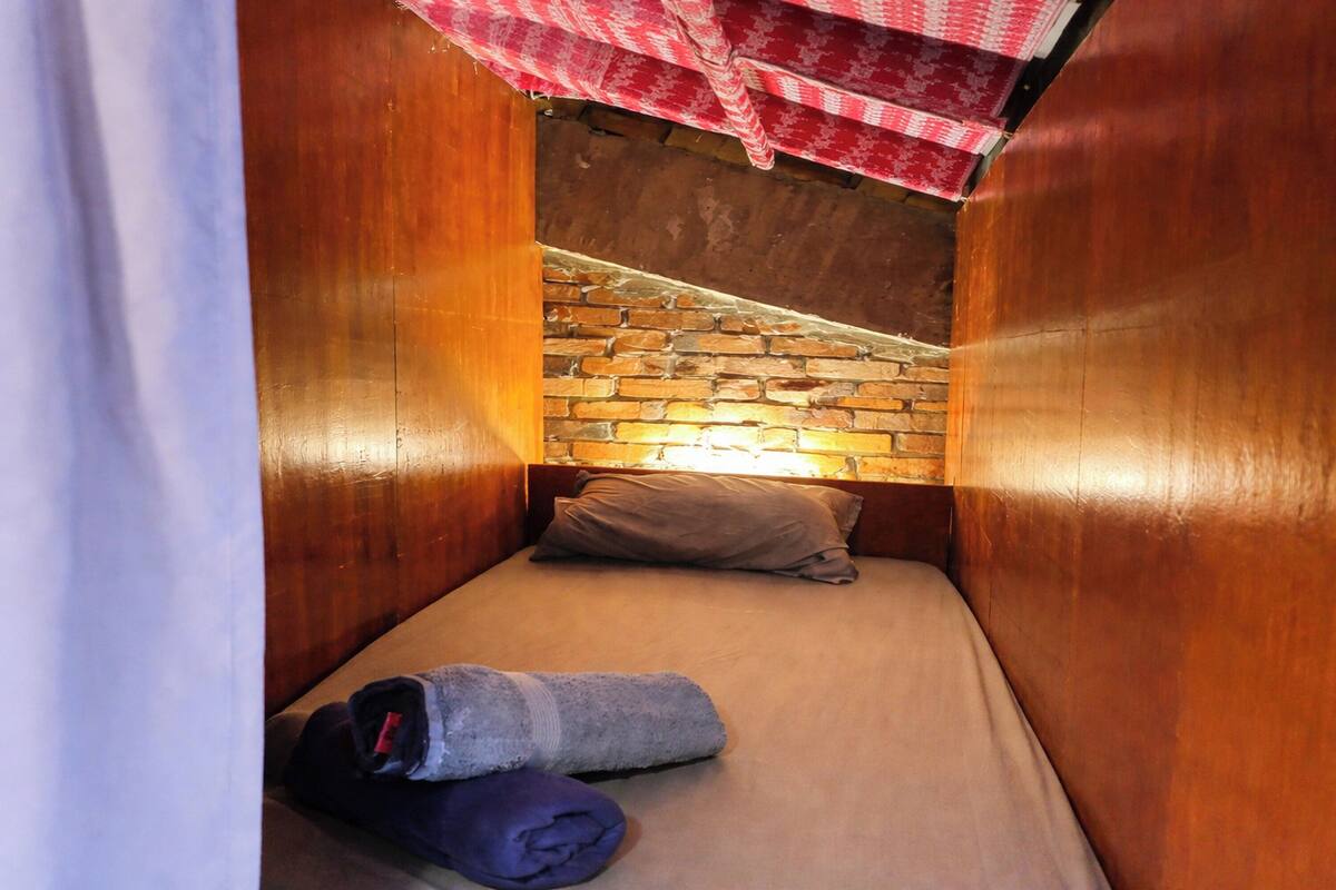 A small, cozy room with a brick wall, wooden paneling, a single bed, and a red and white checkered cloth hanging above.